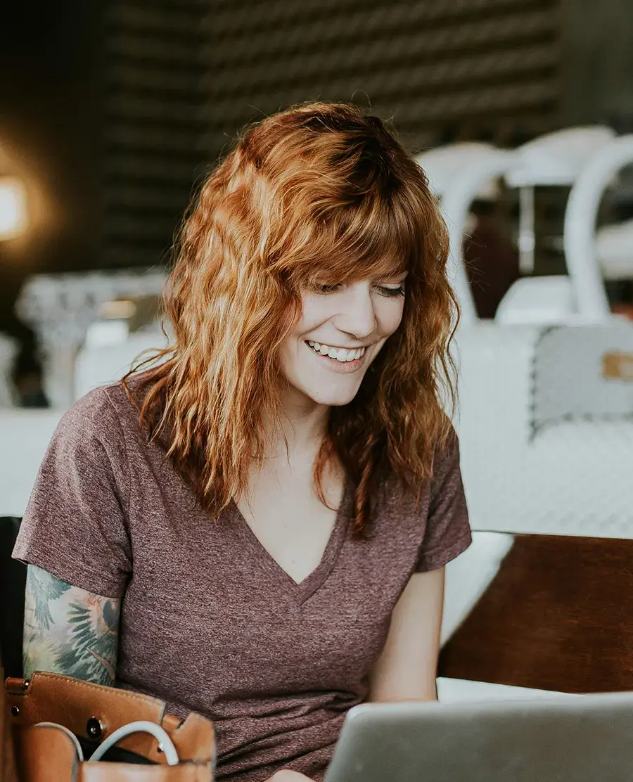 Eine junge Frau mit roten Haaren blickt lachend in ihr Laptop